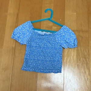 Blå smockad topp med puffärmar - Supersöt blå topp med smockad kropp och korta puffärmar. Toppen har ett vitt prickigt mönster på ärmarna och en croppad passform. Perfekt för dig som gillar en söt och trendig stil med romantiska vibbar.