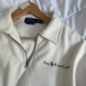 Vit half zip tröja Polo Ralph Lauren - Snygg vit långärmad tröja från Polo Ralph Lauren med relaxed fit. Tröjan har half zip-dragkedja, broderad logga på bröstet och stor känguruficka framtill. Perfekt för en chill och clean look, passar till allt och är superbekväm.