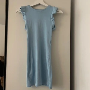 Ljusblå kort klänning med volangärm - Supersöt ljusblå kortklänning med volangdetaljer vid axlarna. Klänningen är ärmlös och har en enkel, rak passform som känns både fräsch och stilren. Perfekt för varma dagar när du vill ha något lätt och bekvämt.