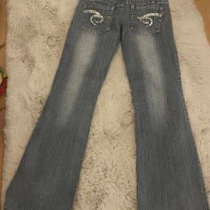 Blå bootcut jeans Bonny Girl strl 27 - Snygga blå bootcut low waist jeans från Bonny Girl med ljusa partier framtill. Bakfickorna har coola broderade detaljer i silver. Klassisk femficksmodell i jeansmaterial, perfekt för dig som gillar en retro vibe. Dem är uppsydda men det är enkelt att ta ner. Svarar på alla frågor eller om nån vill veta mått 