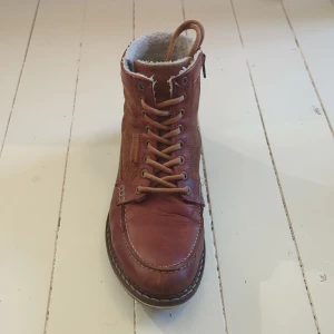 Bruna Björn Borg boots med foder - Säljer ett par snygga bruna boots från Björn Borg i storlek 43. Skorna har snörning, perfekt fungerande dragkedja på sidan och är fodrade med mjukt vitt material för extra värme. Ovandelen är i skinn och mocka, med grov sula som ger bra grepp. Perfekta för kyliga dagar!