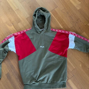 Grön och röd hoodie från FILA - Säljer en cool grön hoodie från FILA med röda och vita detaljer. Tröjan har en justerbar huva och ribbade muddar. Perfekt för en avslappnad stil. Tröjan är även stor i storleken och passar enkelt till storlek M också!