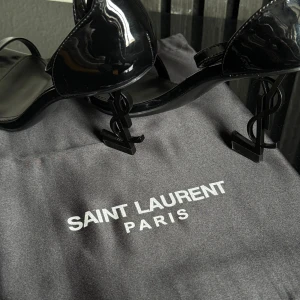 Svarta klackskor från Saint Laurent - Eleganta svarta klackskor från Saint Laurent med en unik klackdesign formad som YSL-logotypen. Skorna har en glansig finish och en smal rem över tårna för en stilren look. Perfekta skor till bröllop, bal, eller andra hög profila event! Alla tillbehör kommer med, även påsen. Observera att boxen har blivit lite smutsig pga flytt men inget som påverkar skon!