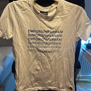 Vit t-shirt från Emporio Armani - Säljer en stilren vit t-shirt från Emporio Armani med blått tryck av märkesnamnet upprepat över bröstet. Perfekt för en casual look. 👕