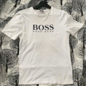 Vit t-shirt från Hugo Boss - Säljer en stilren vit t-shirt från Hugo Boss med deras ikoniska logga tryckt i svart på bröstet. Perfekt för en avslappnad och trendig look.