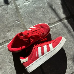 Röda Adidas Campus sneakers - Snygga röda Adidas Campus sneakers med vita ränder och snörning. Perfekta för en casual look med en klassisk design. Skorna har en bekväm passform och är tillverkade i mocka.