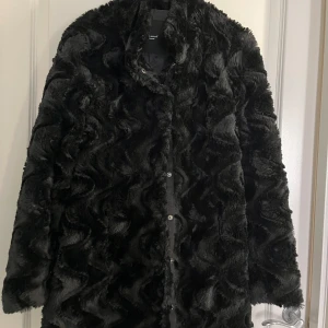 Svart pälsjacka från Vero Moda I M - Snygg svart pälsjacka från Vero Moda med knappar framtill. Jackan har en mjuk och fluffig textur som ger en lyxig känsla. Perfekt för att hålla sig varm och stilren under kyliga dagar.