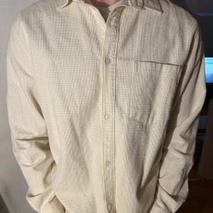 Beige skjorta från Weekday - Säljer en stilren beige skjorta från Weekday. Skjortan har en diskret rutig textur och en bröstficka. Den är långärmad med knappar framtill och en klassisk krage. Perfekt för en avslappnad men ändå snygg look.
