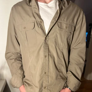 Beige overshirt från H&M - Snygg beige overshirt från H&M med långa ärmar och klassisk krage. Overshirten har två bröstfickor med knappar och axelklaffar för en coolare look. Perfekt för en avslappnad men ändå uppklädd stil.