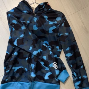 Blå och svart camouflage hoodie - Snygg hoodie i blått och svart camouflage. Den har en dragkedja framtill och långa ärmar. På ärmen finns färgglada bokstäver i orange, blått och rött. Perfekt för en cool och avslappnad stil. Dragkedjan kan dras ändå upp. 