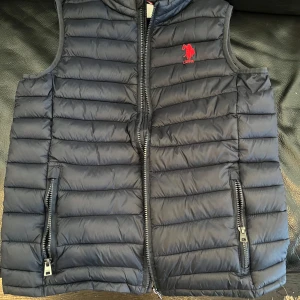 Mörkblå dunväst från U.S. Polo Assn. - Säljer en snygg mörkblå dunväst från U.S. Polo Assn. med dragkedja och två praktiska fickor framtill. Västen har en röd logga på bröstet och är perfekt för kyligare dagar. Passar både till vardags och mer avslappnade tillfällen.