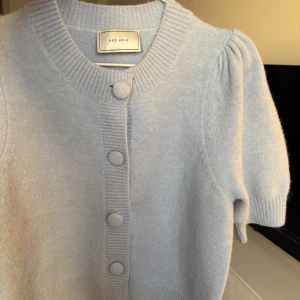 Ljusblå kofta från Neo Noir - Säljer en söt ljusblå kofta från Neo Noir med korta ärmar och knappar framtill. Koftan har en ribbad krage och puffiga ärmar som ger en trendig look. Perfekt för lager-på-lager-stil!