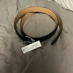 Svart bälte från Como - Snyggt blått bälte från Como med en stilren design. Bältet har en silverfärgad spänne och är tillverkat i ett material som liknar skinn. Perfekt för att ge en elegant touch till din outfit.