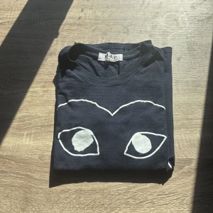 Svart t-shirt från Comme des Garçons - Säljer en svart t-shirt från Comme des Garçons med ett stort vitt hjärta och ögon på framsidan. T-shirten är kortärmad och har en normal passform. Perfekt för en stilren och unik look.