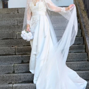 Vit brudklänning med spetsdetaljer - Elegant vit brudklänning med långa spetsärmar och vacker spetsdetaljering på överdelen. Klänningen har en lång släp och en vacker v-ringad design framtill. Baktill finns knappar som ger en klassisk look.