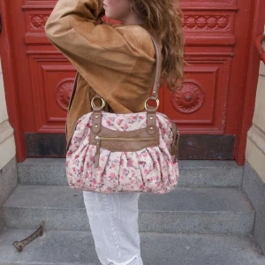 Blommig väska med bruna detaljer - Snygg axelväska med blommigt mönster i rosa och beige. Väskan har bruna skinnliknande detaljer.