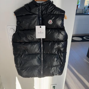 Svart dunväst med luva från Moncler - Snygg svart dunväst från Moncler med dragkedja och hög krage. Perfekt för kyliga dagar. Västen har två praktiska fickor med dragkedja och Monclers logotyp på bröstet. Västen har också en  snygg luva med Moncler märke, knappt använd och etiketterna finns kvar. Pris kan diskuteras!