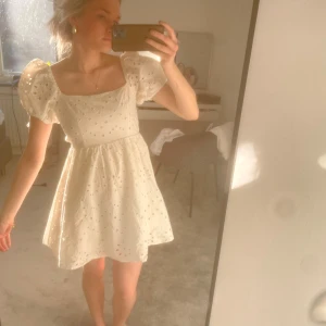 Vit klänning med puffärmar - Charmig vit klänning med puffiga kortärmar och öppen rygg. Klänningen har ett subtilt blommigt mönster och knytning i ryggen. Perfekt för sommarens festligheter.