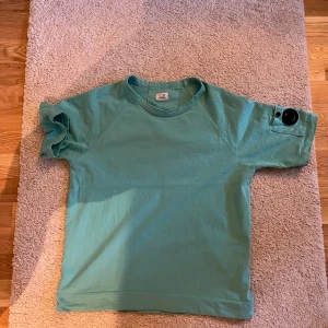 Turkos t-shirt från C.P. Company - Säljer en snygg turkos t-shirt från C.P. Company. Den har korta ärmar och en cool detalj med en knapp och lins på ärmen. Perfekt för en avslappnad stil. 💙väldigt unik bra för sommaren och vid frågor bara skriv. Pris går att pruta!!