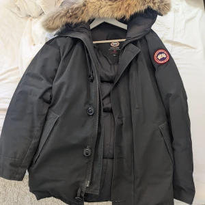 Svart dunjacka från Canada Goose - Säljer en svart dunjacka från Canada Goose med pälsfodrad huva. Jackan har både dragkedja och knappar framtill samt en ikonisk logga på ärmen. Perfekt för kalla vinterdagar.