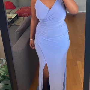 Ljuslila långklänning - Elegant ljuslila långklänning med omlott-design och en smickrande slits. Klänningen har en oneshoulder-stil som ger en modern touch. Perfekt för speciella tillfällen.