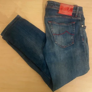 Jacob Cohen jeans  - Snygga blå jeans från Jacob Cohen med röda sömmar och knappgylf. Jeansen har en klassisk femficksdesign och är tillverkade i högkvalitativt denim. Perfekta för en stilren look. De är lite slitna men fixade 