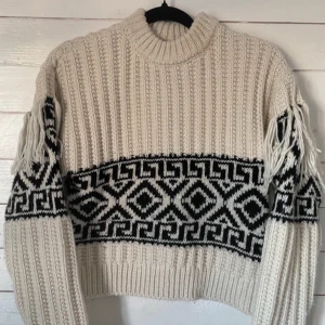 Beige stickad tröja från H&M - Säljer en beige stickad tröja från H&M med svart geometriskt mönster och fransdetaljer på ärmarna. Tröjan är croppad och har en ribbad design med långa ärmar. 