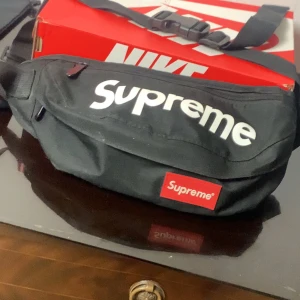 Svart axelväska från Supreme - Snygg svart axelväska från Supreme med justerbar rem och dragkedja. Perfekt för att bära dina nödvändigheter med stil. Den har en tydlig Supreme-logga i vitt på framsidan och en röd etikett med loggan.