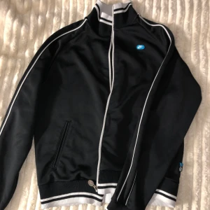 Svart vindjacka med vita detaljer - Snygg svart vindjacka från Nike med vita ränder längs ärmarna och dragkedja framtill. Jackan har ribbade muddar och en liten logotyp på bröstet. Perfekt för en sportig look!
