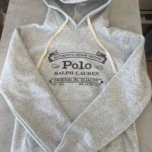Grå hoodie från Ralph Lauren - Säljer en snygg grå hoodie från Ralph Lauren med tryck på framsidan. Den har en klassisk design med dragsko i huvan och ribbade muddar vid ärmslut och nederkant. Perfekt för en avslappnad stil.
