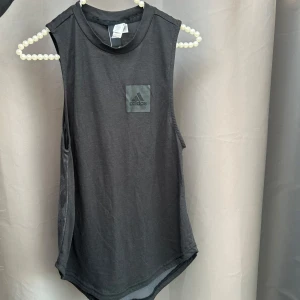 Svart ärmlös topp från Adidas - Säljer en svart ärmlös topp från Adidas med logga på bröstet. Perfekt för träning eller en avslappnad dag. Tillverkad i mjukt och bekvämt material.🩶🖤