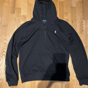 Svart hoodie från Polo Ralph Lauren - Säljer en stilren svart hoodie från Polo Ralph Lauren med dragkedja och broderad logga på bröstet. Den har en justerbar huva och långa ärmar. Perfekt för en avslappnad stil. Har krympt lite så den passar som M