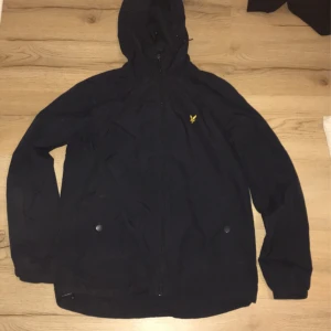Svart jacka med huva från Lyle & Scott - Säljer en svart jacka med huva från Lyle & Scott. Jackan har en dragkedja framtill och är dekorerad med ett gult broderat emblem på bröstet. Perfekt för kyliga dagar.