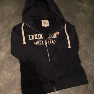 Mörkblå hoodie från Lexington - Säljer en snygg mörkblå hoodie från Lexington med dragkedja och tryck framtill. Den har en huva med vita snören och ribbade muddar vid ärmslut och nederkant. Perfekt för en avslappnad stil.