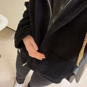 Svart teddyjacka med dragkedja - Mysig svart teddyjacka med dragkedja framtill. Perfekt för kyliga dagar med sin fluffiga och varma design. Jackan har långa ärmar och en avslappnad passform.