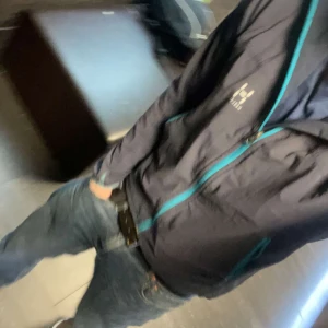 Grå vindjacka från Haglöfs - Säljer en grå vindjacka från Haglöfs med turkosa dragkedjor. Jackan har en justerbar huva och långa ärmar. Perfekt för blåsiga dagar. Ny skick! 