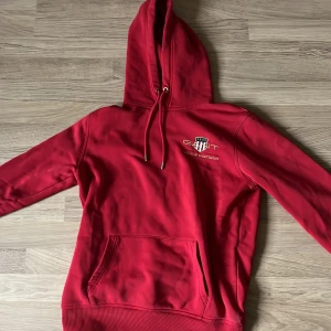 Röd hoodie från GANT - Säljer en snygg röd hoodie från GANT med en klassisk känguruficka och justerbar huva. Perfekt för en avslappnad stil. Den har en liten logotyp på bröstet.