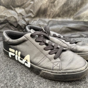 Svarta sneakers från FILA - Svarta sneakers från FILA med vit logga på sidan. Skorna har snörning och är tillverkade i syntetmaterial med platt sula och rund tå. Perfekta för en stilren och sportig look.