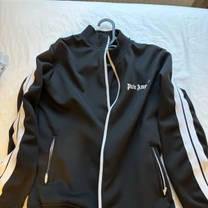 Svarta träningsbyxor från Palm Angels - Svarta träningsbyxor från Palm Angels med vita sidoränder och logga på vänster lår. Byxorna har resår i midjan och raka ben. Perfekta för en sportig look. Har används ett par gånger och däremot tagit väl hand om dressen. Forrftaraden helt ren och fräsch. Vill man köpa enskild fungerar det utmärkt. Man får även med dressens påsar 