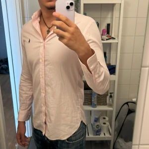 Ljusrosa skjorta från Ralph Lauren - Säljer en ljusrosa skjorta från Ralph Lauren med klassisk krage och blå broderad logga på bröstet. Skjortan har långa ärmar och knäppning framtill. 