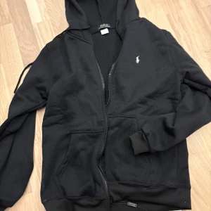 Svart hoodie från Polo Ralph Lauren - Svart hoodie från Polo Ralph Lauren med dragkedja framtill och klassisk logga broderad på bröstet. Tröjan har huva med dragsko och två fickor framtill. Perfekt för en avslappnad stil.