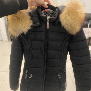 Svart pufferjacka med päls från Garoff - Säljer en svart pufferjacka från Garoff med stor fluffig päls runt huvan. Jackan har dragkedja framtill och två snedställda fickor med dragkedja. Perfekt för kyliga dagar och riktigt snygg siluett.