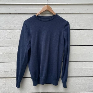 Mörkblå stickad tröja - En stilren mörkblå stickad tröja med rund halsringning och långa ärmar. Tröjan har ribbade muddar vid ärmslut och nederkant, vilket ger en klassisk och enkel look. Perfekt att matcha med jeans eller chinos.