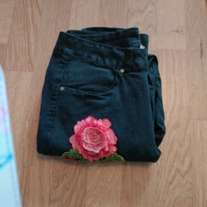 Svarta jeans med broderad ros - Svarta jeans med snygg broderad rosa ros och gröna blad på ena benet. Klassisk femficksmodell med raka ben och normal passform. Perfekta för dig som vill sticka ut lite extra med detaljer.