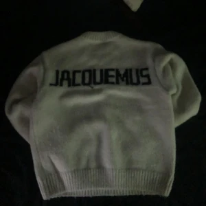 Vit stickad tröja från Jacquemus - Vit stickad tröja från Jacquemus med rund halsringning och svart Jacquemus-logga på ryggen. Tröjan har ribbade muddar vid ärmslut och nederkant. Perfekt för en stilren och trendig look. 