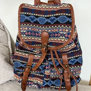 Ryggsäck  - Färgglad ryggsäck med etniskt mönster i tyg och detaljer i brunt konstläder. Ryggsäcken har ett lock som stängs med ett spänne, samt dragsko. Justerbara axelremmar och ett litet fack med dragkedja på baksidan. Inuti finns ett rymligt fack och ett mindre fack med dragkedja. Ryggsäcken är i fint skick.   Mått: Höjd ca 35 cm Bredd ca 30 cm Djup ca 18 cm