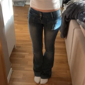 Blå bootcut jeans med dubbla knappar - Säljer ett par blå bootcut jeans med dubbla knappar i midjan och stora fickor fram och bak. Jeansen har en klassisk tvätt och markerade sömmar. Perfekta för dig som gillar en retroinspirerad look. Midjemåttet rätt över är 37 cm och innerbenslängden är ca 80 cm
