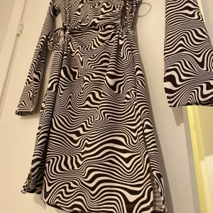 Svartvit mönstrad omlottklänning - Säljer en svartvit klänning med vågigt, randigt mönster. Klänningen har långa ärmar, knytband i midjan och vid sidorna samt en lätt figurnära passform. Materialet känns mjukt och stretchigt, perfekt för dig som gillar unika mönster.