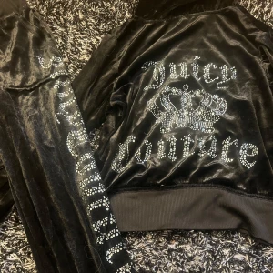 Svart velour tracksuit från Juicy Couture - Juicy Couture svart velour-set med hoodie och byxor. Hoodien har dragkedja, fickor och strassdetaljer med logga och krona på ryggen. Byxorna har strasslogga längs benet och fickor med knappar. Ej äkta 