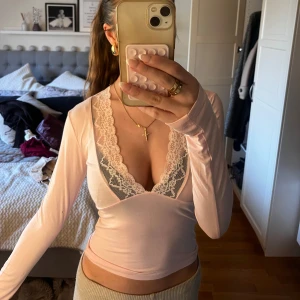 Ljusrosa långärmad topp med spetsdetaljer - Säljer en ljusrosa långärmad topp med djup v-ringning och fina spetsdetaljer längs urringningen. Jätte fin med en ljusare Bh